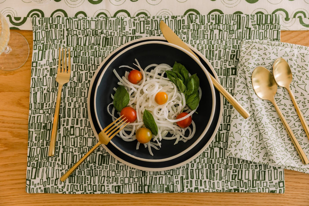 Green and white printed linen napkins and placemat with a matching table runner, brass flatware, and a salad of noodles and tomatoes